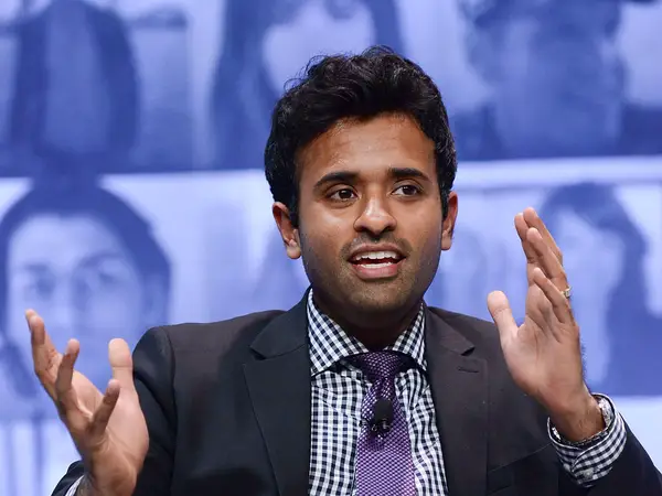 Photograph of a man in his 20s who has heritage from India with medium skin tone and crew cut black hair. He is wearing a black suit, blue and white gingham button up and purple tie. He is seated on stage with a screen comprised of faces behind him. He is gesturing with his hands. 