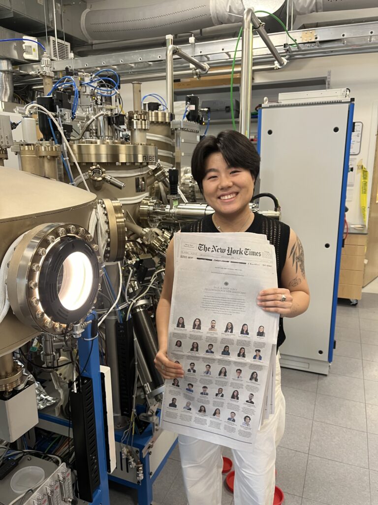 Photograph of a non-binary person in their 20s who has heritage from China with light skin tone and black pixie hair with curtain bangs. They are standing in a lab surrounded by technical equipment and holding the New York Times.