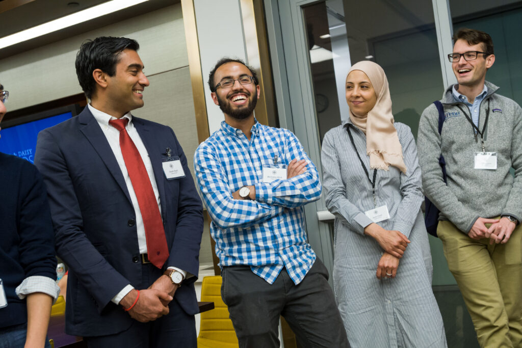 Photograph of three people standing, the one in the middle is a man in his 20s with medium skin tone, black short hair and a black beard - he is smiling. There is a man on the left in his 20s with medium skin tone and short black hair, on the right a woman in her 20s with light-medium skin tone a cream colored hijab and grey dress - she is smiling. They are both looking at the man in the middle. 