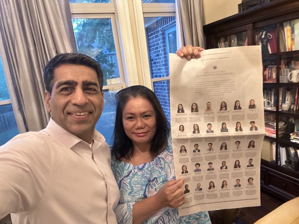 Parents of a fellow stand stand holding the New York Times, there is a window behind them showing a red brick house.