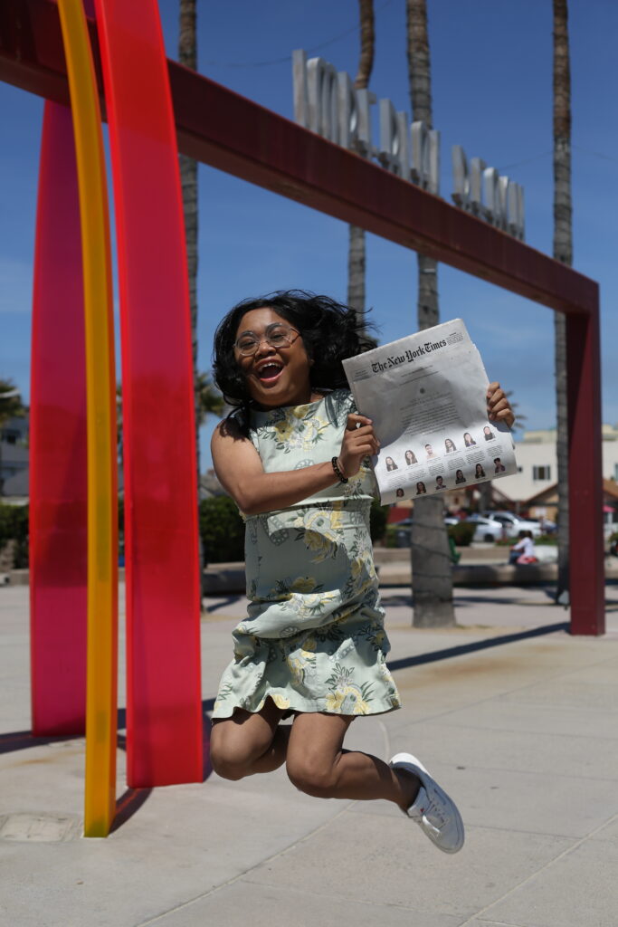 Photograph of a woman in her 20s who has heritage from Philippines with light-medium skin tone and long black hair. She is mid-jump in excitement holding the New York Times.