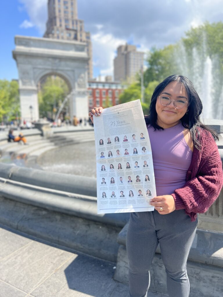 Photograph of a woman in her 20s who has heritage from Honduras with light skin tone and long black hair. She is standing in front of a fountain and in the background the Washington Square Arch can be seen. 