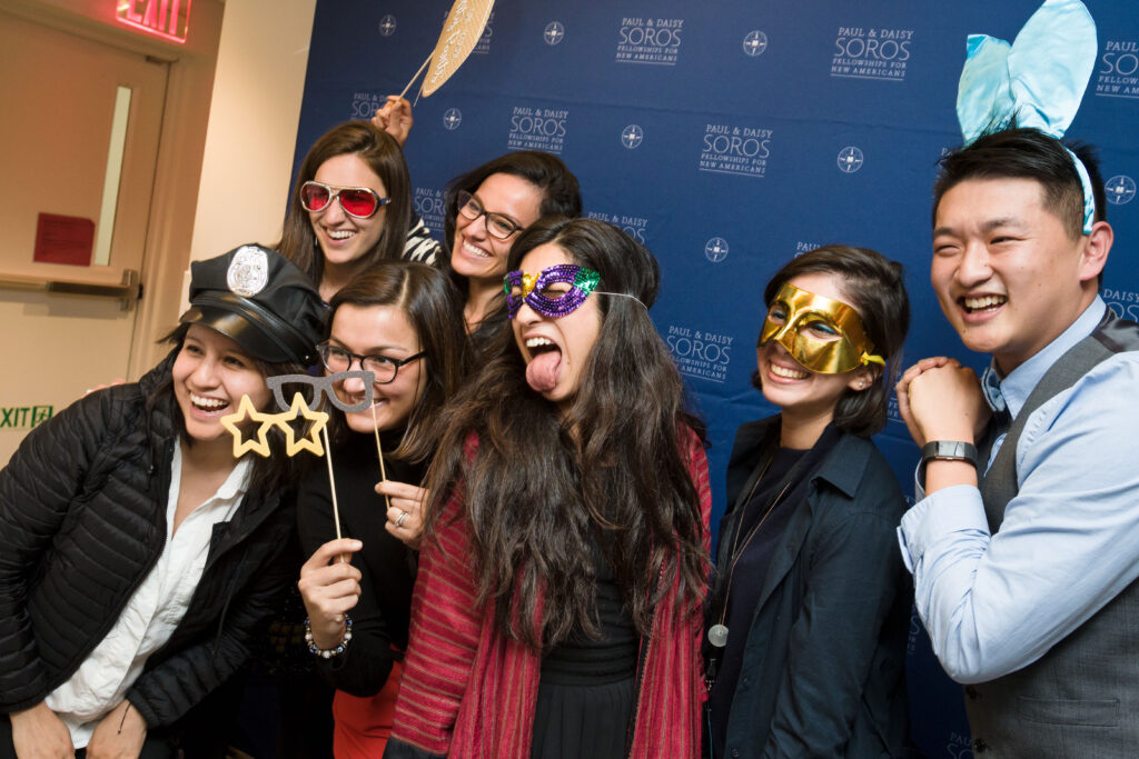 Photograph of seven people in their 20s of varying heritages, in a makeshift photo booth, they are standing in front of a Paul & Daisy Soros logo navy step and repeat. They are all wearing various props (masquerade masks, giant bow, sunglasses, etc.) and smiling or making a silly face at the camera.