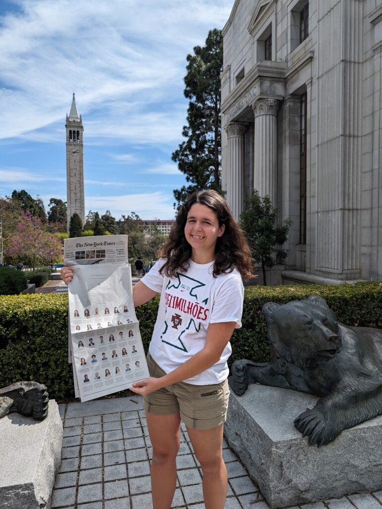 Photograph of a woman in her 20s who has heritage from Portugal who has light skin tone and shoulder length dark brown hair. She is standing in front of a grey stone building with columns. 