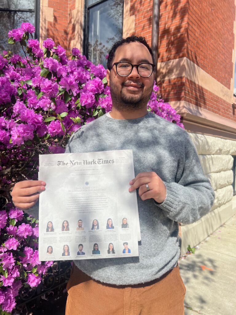 Photograph of a man in his 20s who has heritage from Colombia with light skin tone, short black hair, short beard and glasses. He is standing in front of a red brick building and bright pink flowers.