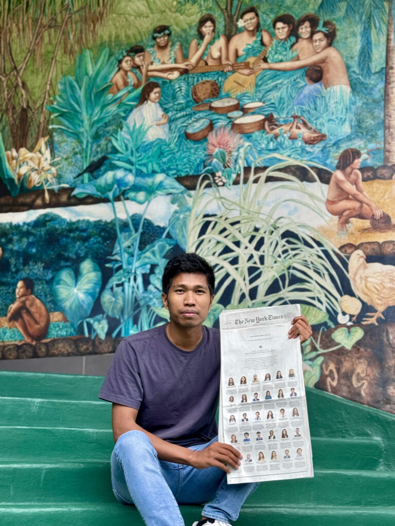 Photograph of a man in his 20s who has heritage from Myanmar with medium skin tone and short black hair. He is sitting on a green bench in front of a mural depicting people in a tropical forest doing various activities. 