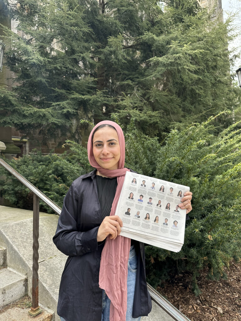 Photograph of a woman in her 20s who has heritage from Palestine with light skin tone. She is wearing a pink hijab. She is standing on a staircase with trees behind her.