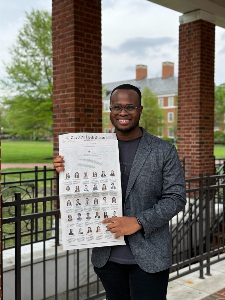 Photograph of a man in his 20s who has heritage from Haiti with dark skin tone and buzzed black hair. He is standing under the overhang of a red brick building looking out on trees and green grass. 