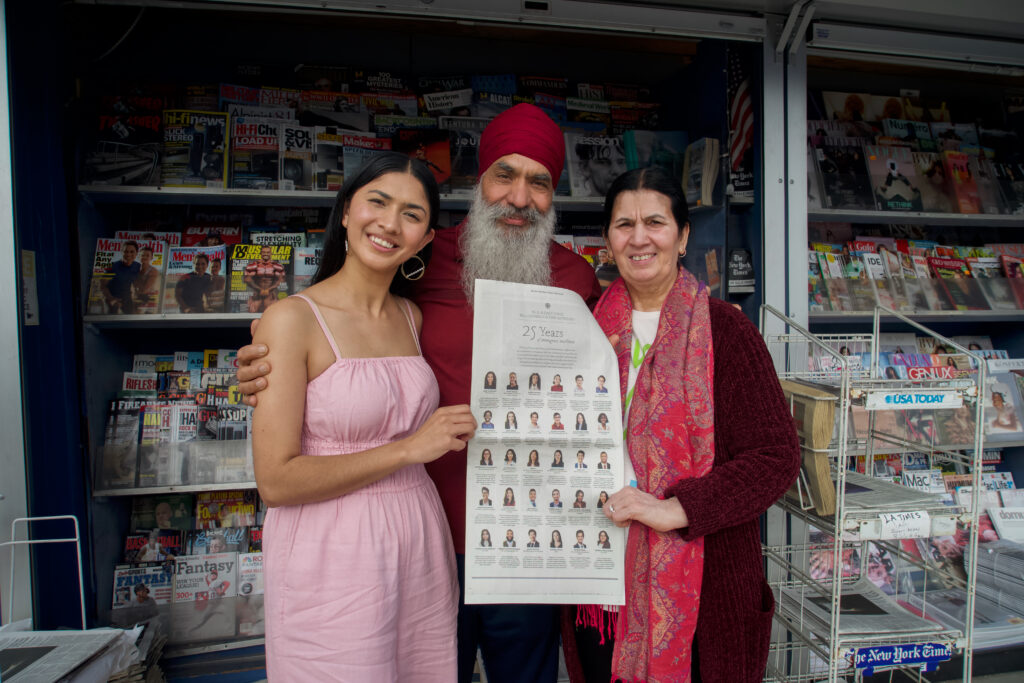 Photograph of a woman in her 20s who has heritage from India with light-medium skin tone and long straight black hair. She is standing in front of a newsstand with her parents. 