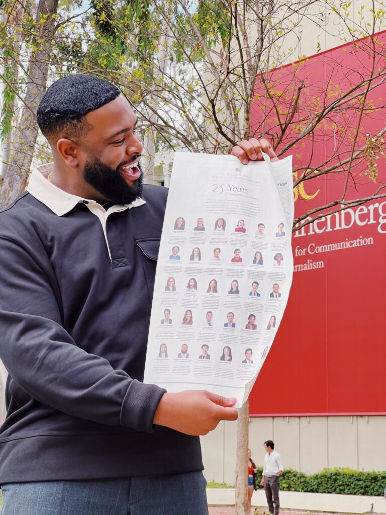 Photograph of a man in his 20s who has heritage from Jamaica with medium skin tone, short black hair and a medium length beard. He is standing in front of a tree, and a red sign can be seen in the background.