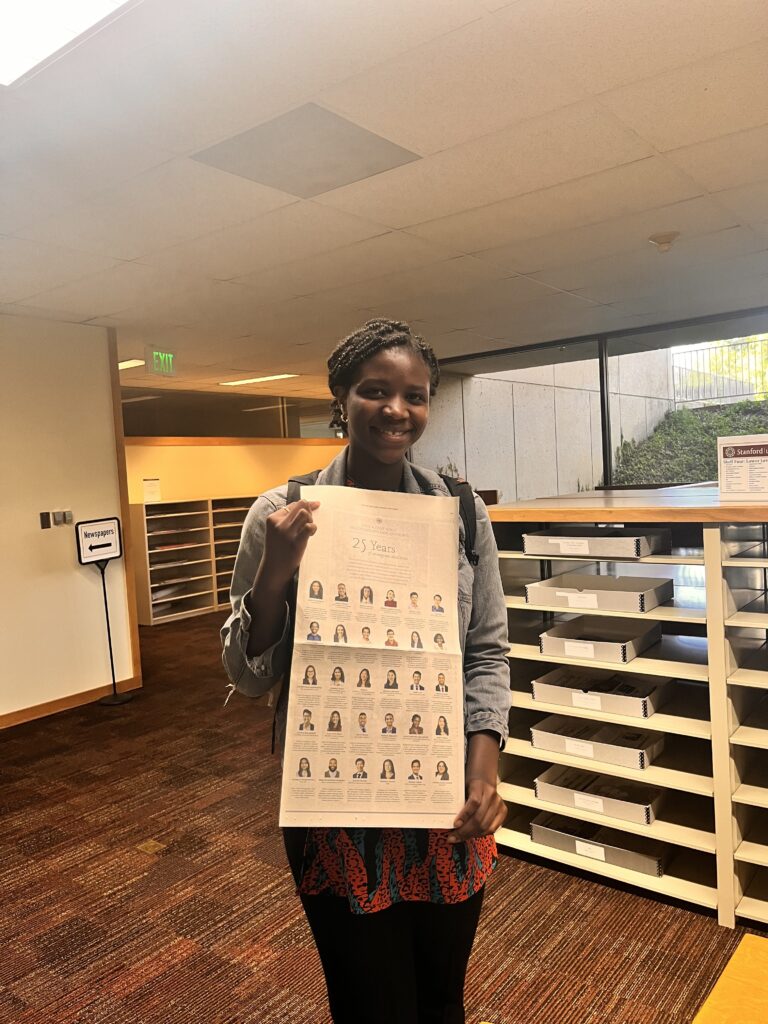 Photograph of a woman in her 20s who has heritage from Nigeria with dark skin tone and black hair braided and pulled back. She is standing in a room with a lot of thin shelves full of boxes. 