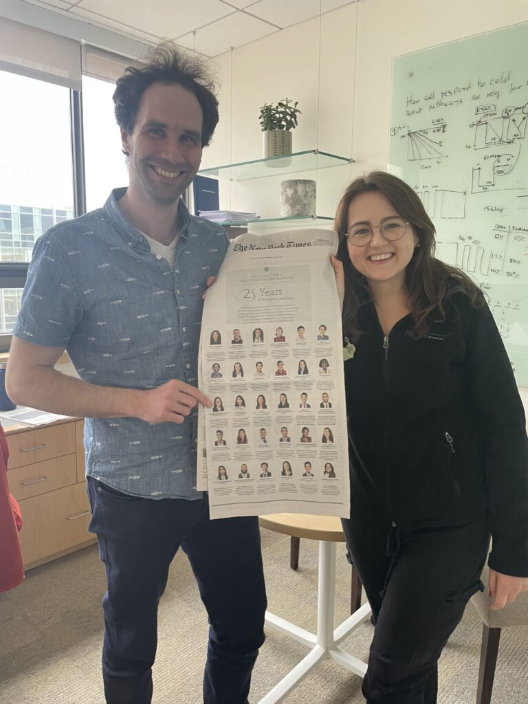 Photograph of a woman in her 20s who has heritage from Poland with light skin tone and long brown hair. She is holding the New York Times with a man. They are standing in a university building with a dry erase board and desk in the background.