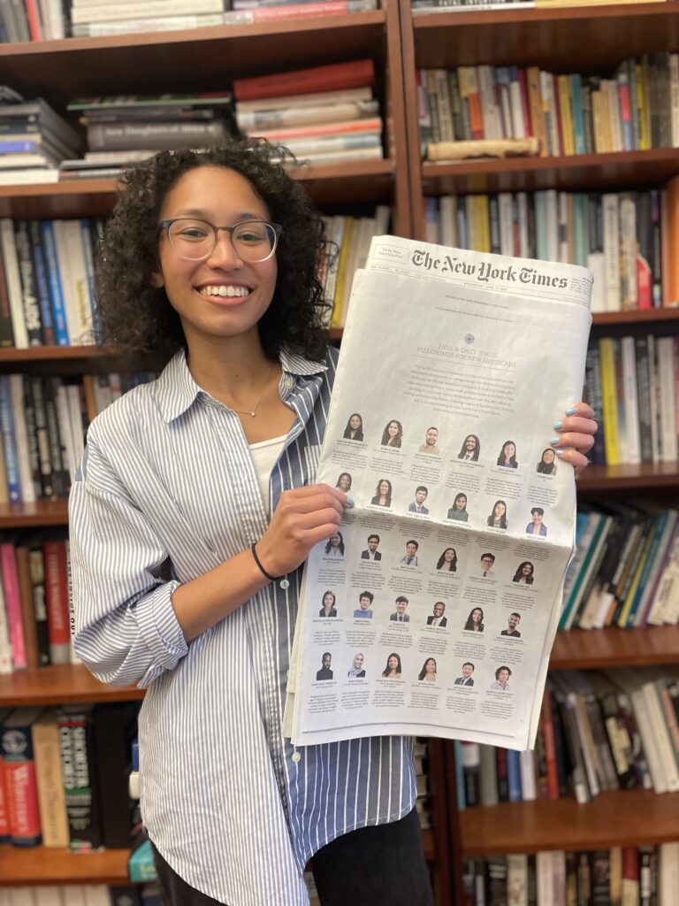 Photograph of a woman in her 20s who has heritage from Haiti and the Philippines with light-medium skin tone and curly shoulder length black hair. She is standing in front of a tall bookshelf, filled with books.