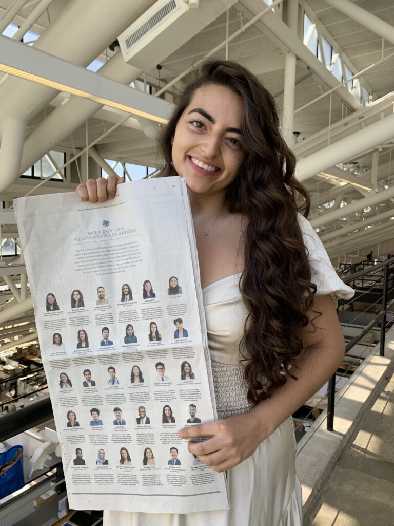 Photograph of a woman in her 20s who has heritage from Mexico with light skin tone and long curled dark brown hair. She is standing in a building with a lot of exposed duct work painted white.