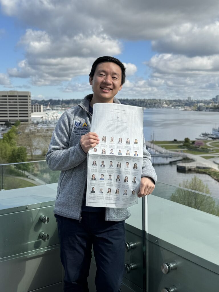 Photograph of a man in his 20s who has heritage from China with light skin tone and short black hair. He is standing on a rooftop overlooking a body of water.