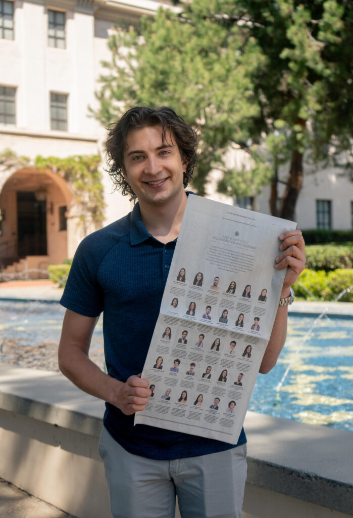 Photograph of a man in his 20s who has heritage from Russia with light skin tone and shaggy brown hair. He is standing in front of a fountain with a building in the background and trees. 