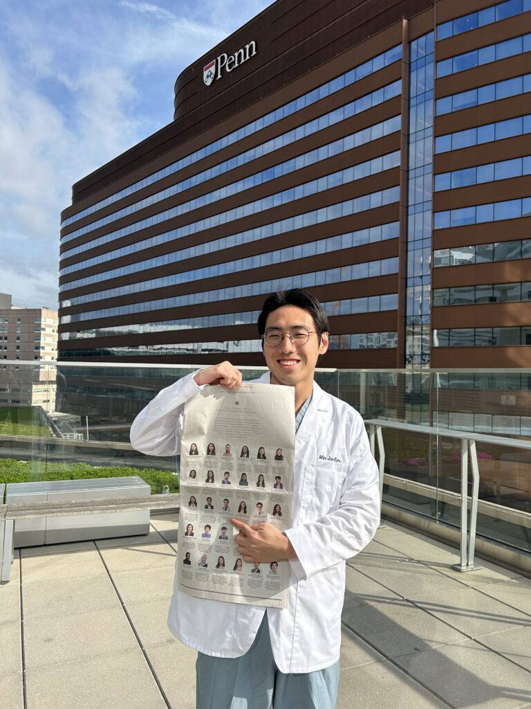 Photograph of a man in his 20s who has heritage from South Korea with light skin tone and short black hair. He stands in front of a large building with many windows that says "Penn" across the top.