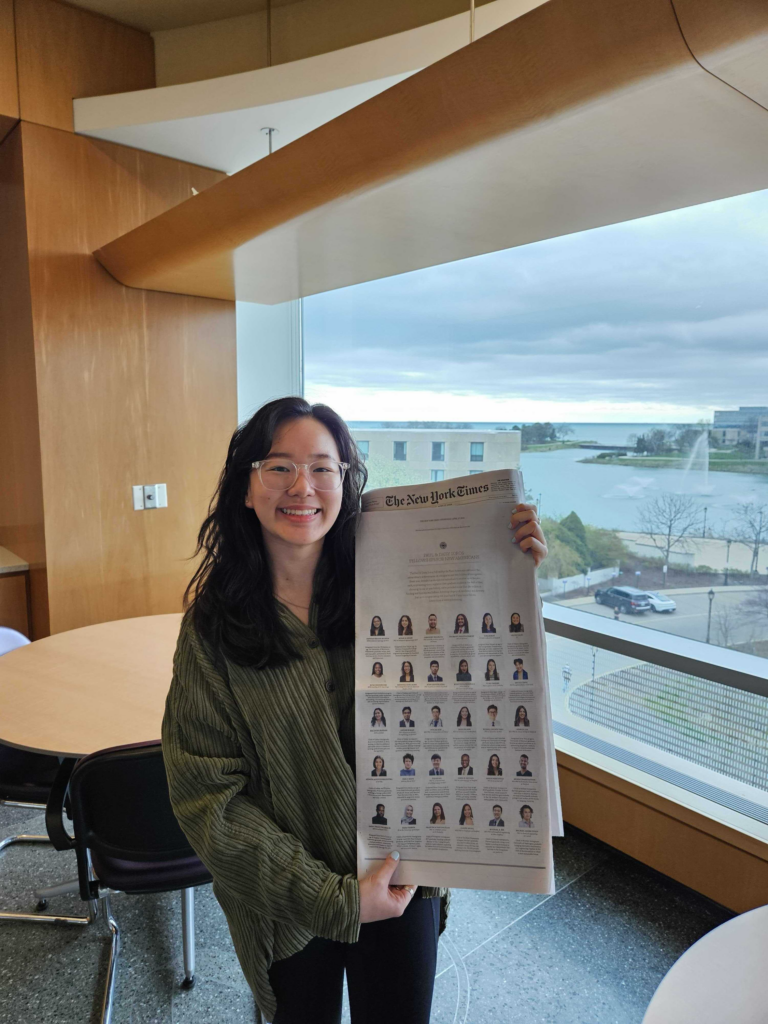 Photograph of a woman in her 20s who has heritage from South Korea with light skin tone and long black hair. She is standing in front of a window overlooking a body of water, there is light wood around her inside. 