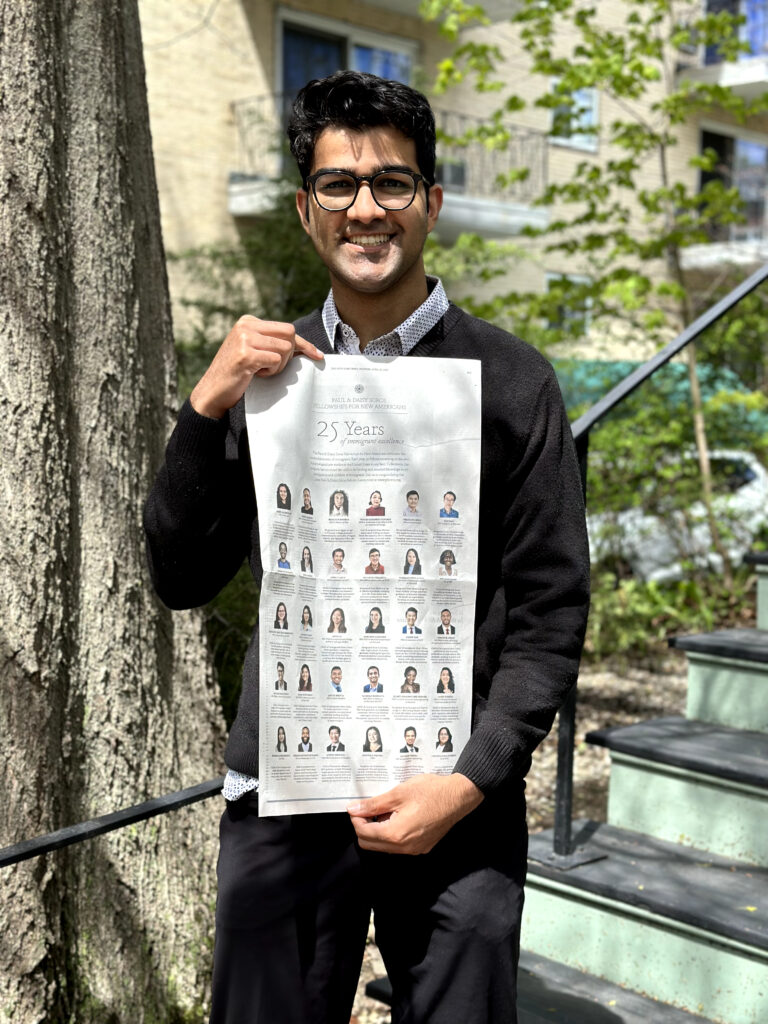 Photograph of a man in his 20s who has heritage from India with medium-light skin tone and short black hair. He is standing at the bottom of a staircase, with a tree trunk to his right. There are more trees and another building in the background.