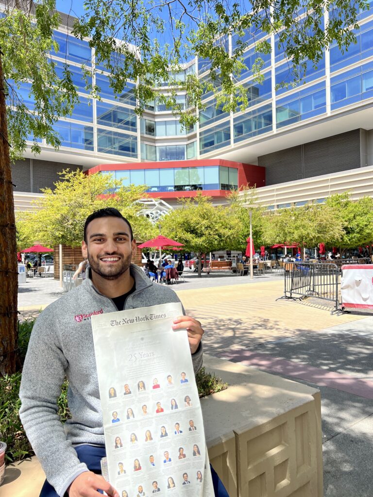 Photograph of a man in his 20s who has heritage from India with light skin tone, short black hair and a short beard. He is standing in a courtyard in front of a large building. 