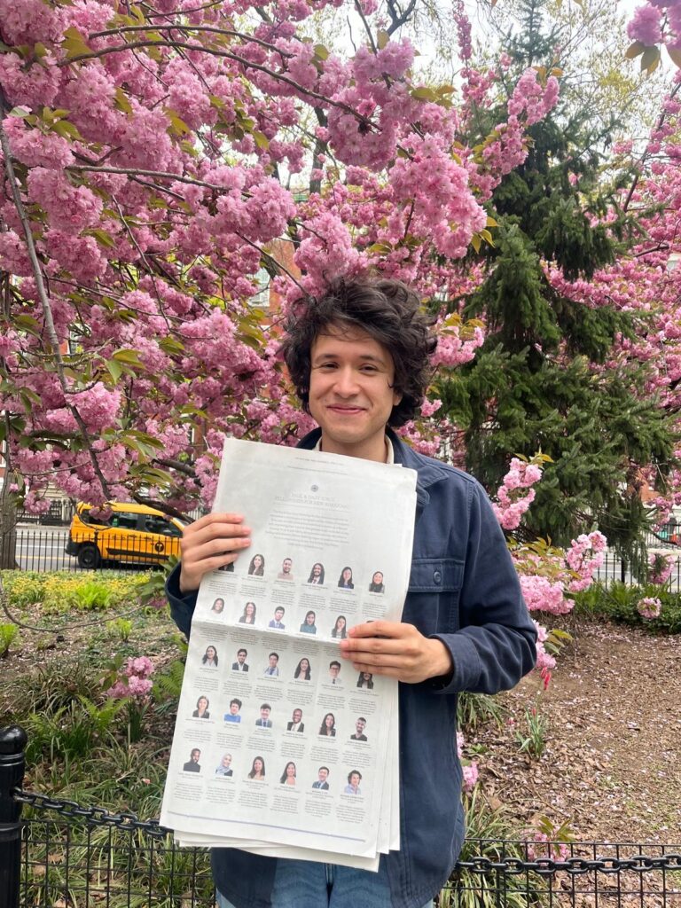 Photograph of a man in his 20s with heritage from Mexico who has light skin tone and shaggy dark brown hair. He is standing in front of cherry blossom trees and holding a New York Times. 