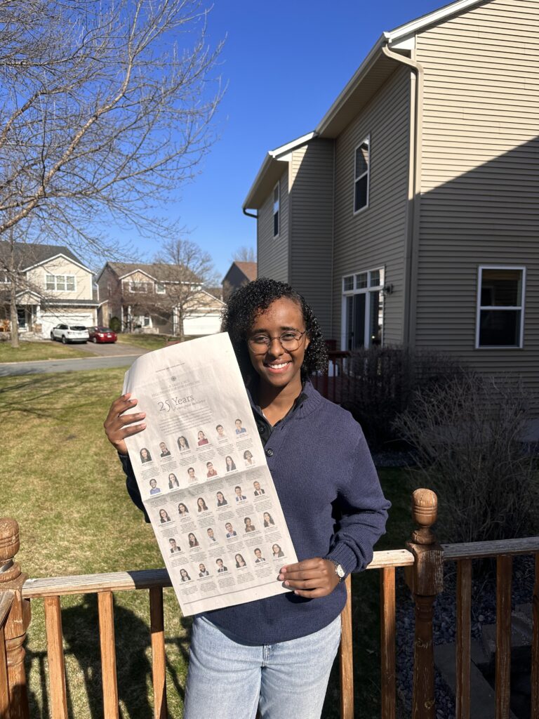 Photograph of a woman in her 20s who has heritage from Somalia with medium-dark skin tone with curly chin length black hair. She is standing in the yard of a suburban house, with more houses and yards in the background. 