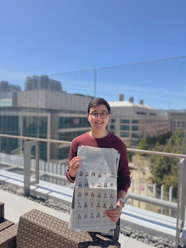 Photograph of a man in his 20s who has heritage from Mexico with light skin tone and short black hair. He is standing outside, with a glass wall behind him, other tall buildings and the tops of chairs can be seen. The sky is blue.