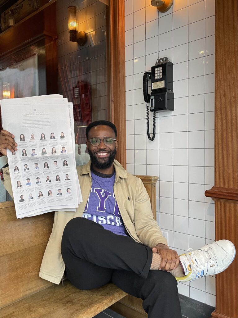 Photograph of a man in his 20s who has heritage from Sierra Leone with dark skin tone, short black hair and short beard. He is sitting on a bench outside a store front, there is a white tile wall next to him with a vintage pay phone.