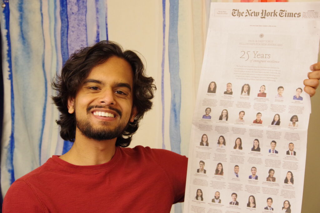 Photograph of a man in his 20s who has heritage from India with light-medium skin tone and shaggy and curly dark brown hair. He is in front of a wall with loose blue stripes. 