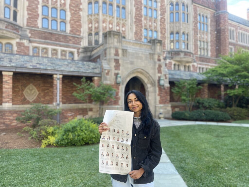 Photograph of a woman in her 20s who has heritage from India with medium skin tone and long black straight hair. She is standing in front of a large stone and red brick building. 