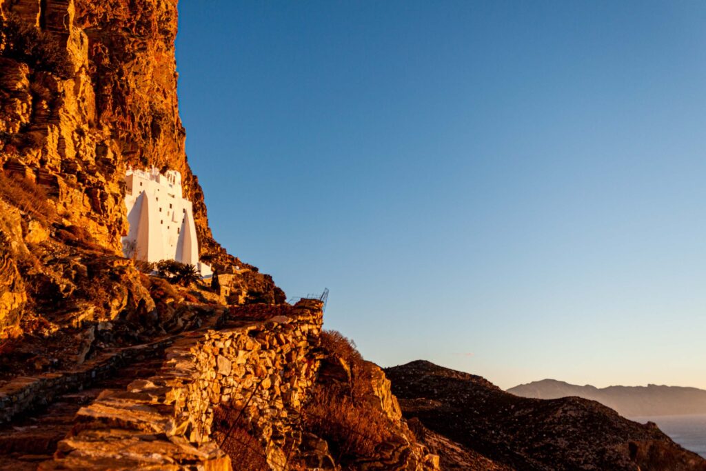 Photograph of a white building built into the side of a rocky mountain. Additional mountains can be seen in the background and the sky is a cloudless blue. 