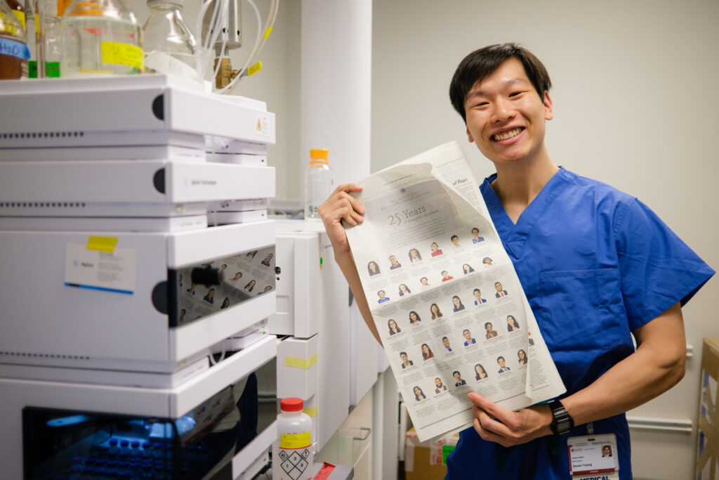 Photograph of a man in his 20s who has heritage from Vietnam with light skin tone and black hair. He is standing in a room that appears to be a lab. 