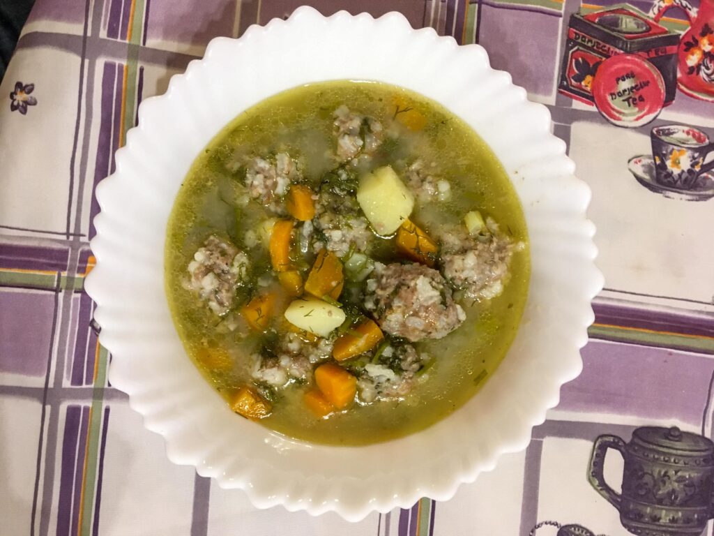 Photograph of a bowl of soup, meatballs, carrots, potatoes and herbs can be seen. The tablecloth is multi-colored, mostly white and purple tartan pattern. 