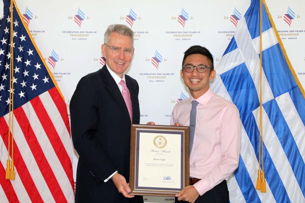 Photograph of a man in his 50s and a man in his 20s, they are posing and holding a framed document together. They stand in front of a step & repeat, an American flag and a Greek flag.