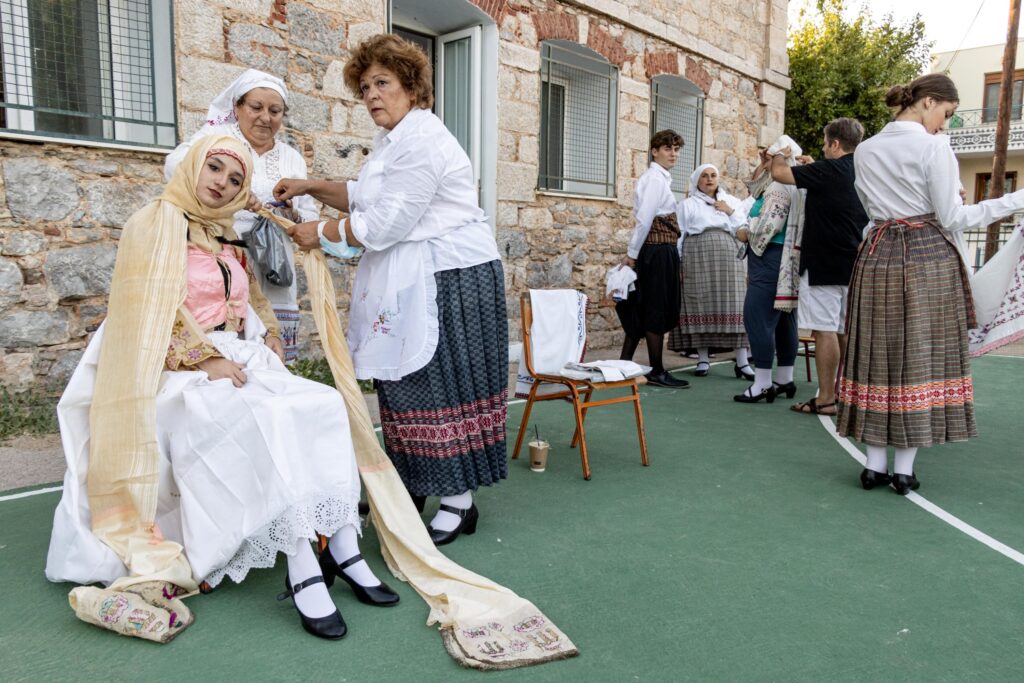 Photograph of several women getting dressed in traditional clothing. Some women are looking at the camera, They are standing outside an old stone building.