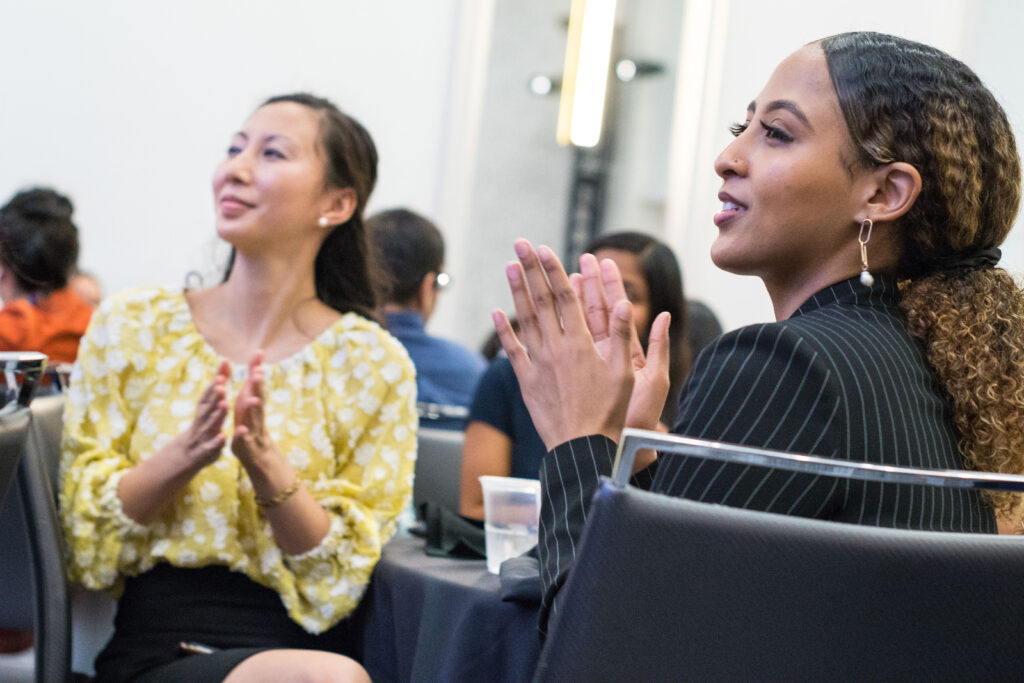 Photograph of two women sitting at a table, looking off camera left. They are both mid-clap. The woman in focus and closer to the camera is in her 20s with medium skin tone and her black wavy hair is pulled back. She is dressed in a suit. The other fully visible woman is in her 20s with light skin tone, long dark hair pulled half back. Other people can be seen sitting nearby.