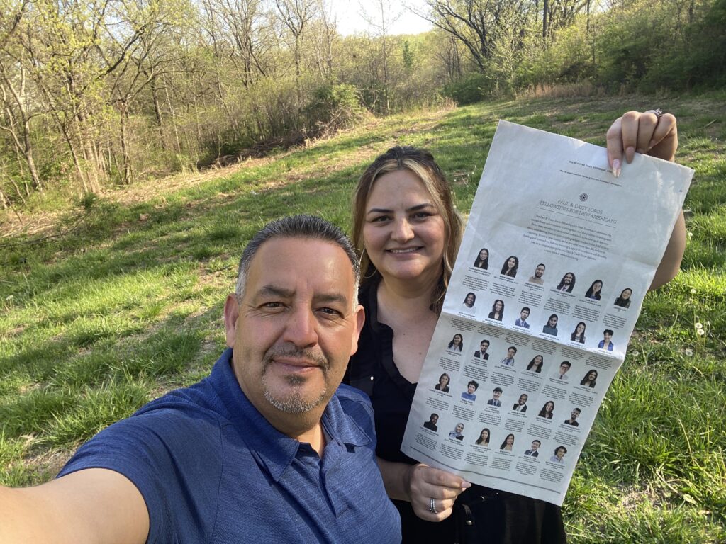 Parents of a fellow stand in a field holding the New York Times and smiling at the camera. 