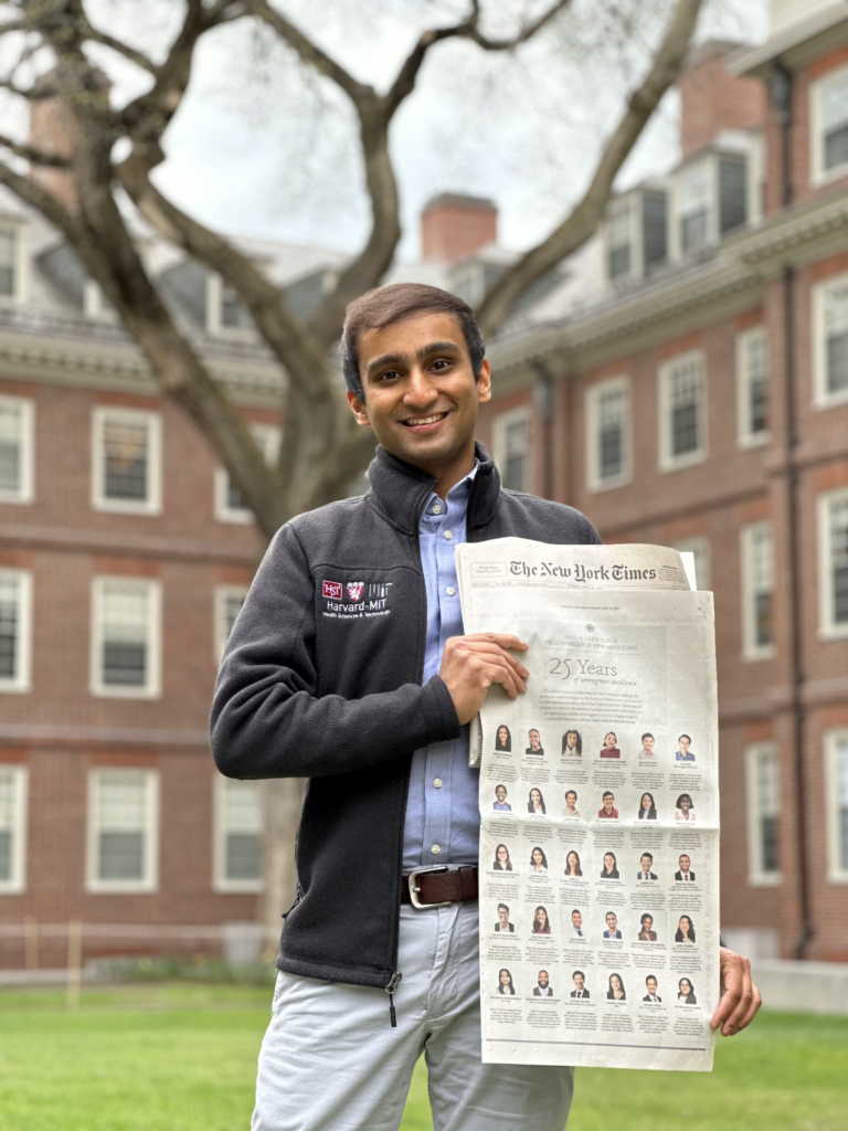 Photograph of a man in his 20s who has heritage from India, with medium skin tone and dark brown short hair. He is standing on a lawn with a bare tree behind him, and a large red brick building behind that. 