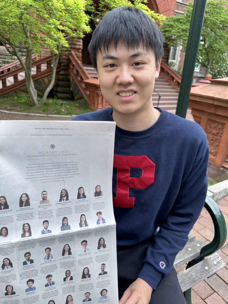 Photograph of a man in his 20s who has heritage from China with light skin tone and black hair. He is standing in front of a red brick building.