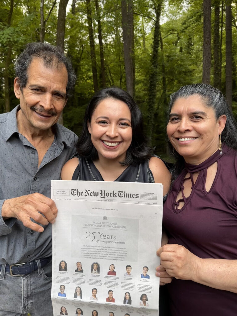 Photograph of a woman in her 20s who has heritage from Mexico, with light skin tone and shoulder length black hair. Her parents stand on either side of her, a forest can be seen behind them. 