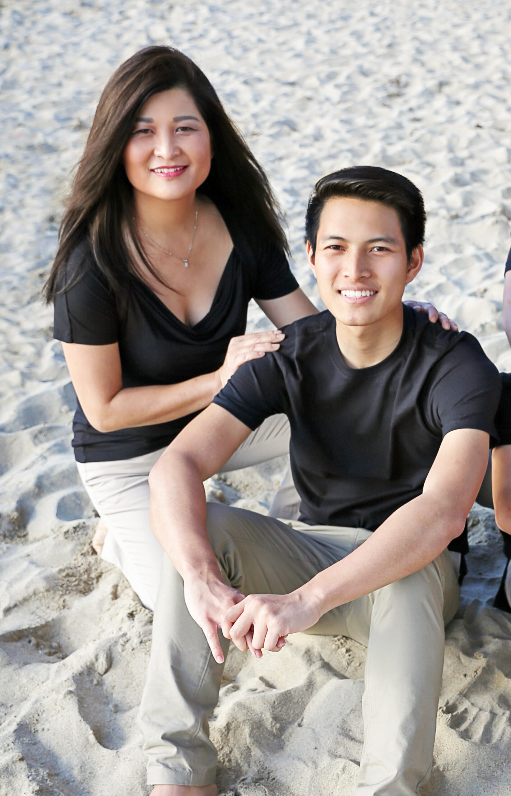 Photograph of two people both wearing khakis and black t-shirts. They are sitting on sand. The man is in his 20s who has heritage from Vietnam with light skin tone and black combed over hair. 