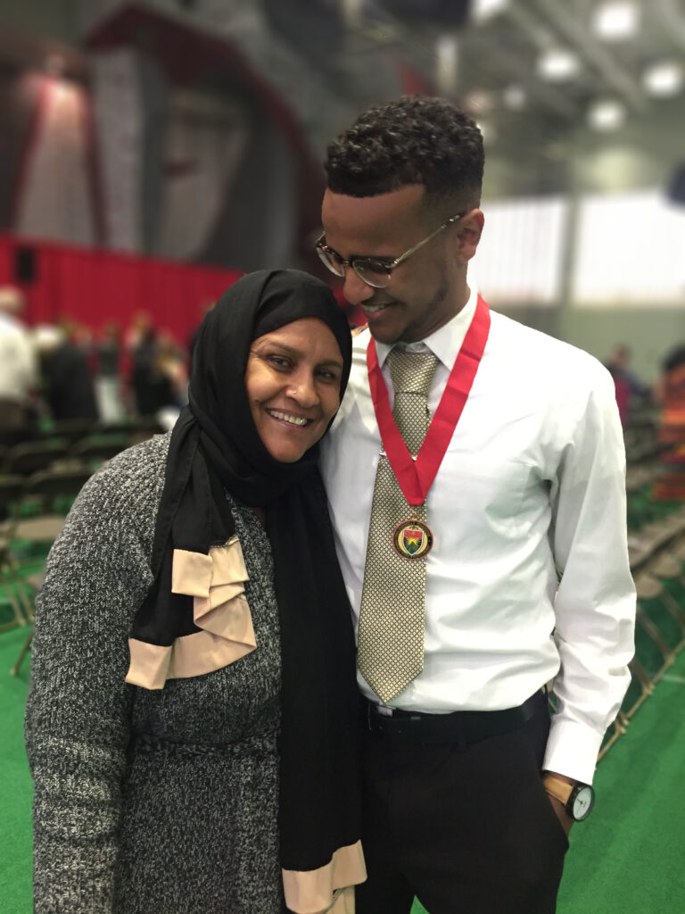 Photograph of a man in his 20s with medium-dark skin tone and short black hair, wearing a white button up, gold tie black pants and glasses. He has his arm around and is looking down at a woman to his right. She is middle-aged and wearing a black and tan hijab with a black and white heathered sweater. 