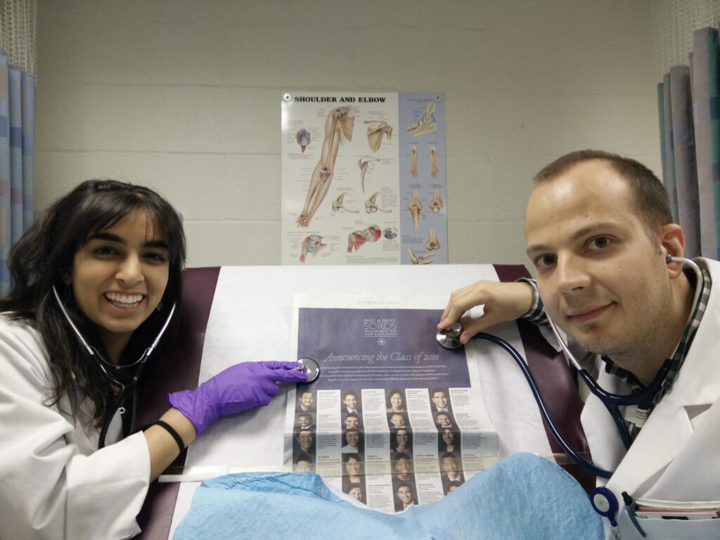 Photograph of two people in a doctor's examination room, they are both wearing doctor's white coats. The woman on the left is in her 20s with heritage from India with medium skin tone and long black hair with bangs. The man on the right is in his 20s with light skin tone and buzzed blonde hair. They are pretending to "examine" with stethoscopes the New York Times that is laid out on the table in front of them. 