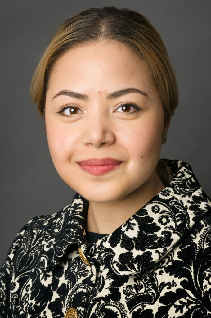 Headshot of a woman in her 20s who has heritage from Libya with light skin tone and dyed dark blonde hair. She is wearing a black and white floral patterned jacket with gold buttons. She is looking at the camera. 