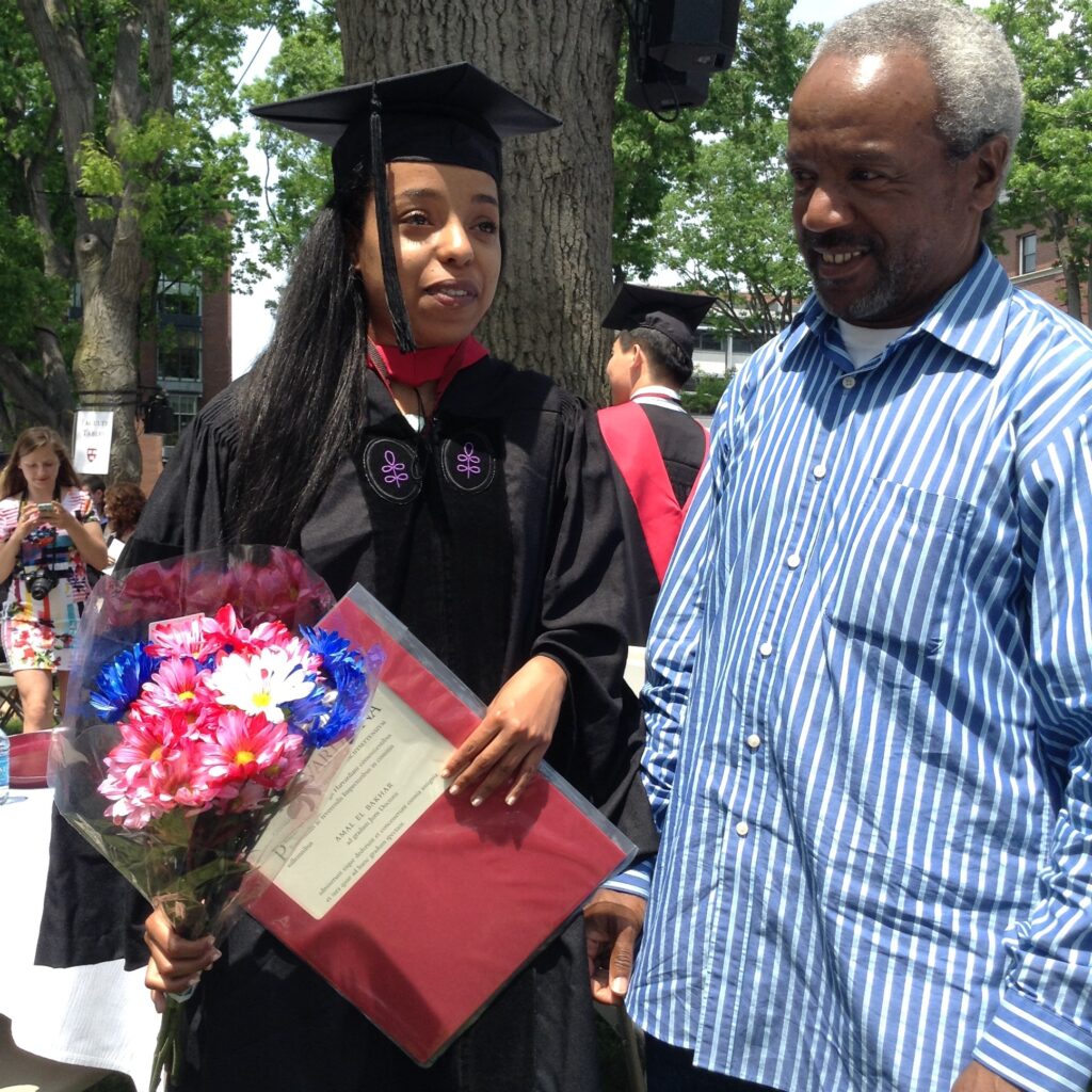 Photograph of two people. A woman in her 20s on the left who has heritage from Morocco with medium skin tone and long black straight hair wearing graduation gown and cap, holding a diploma and bouquet of flowers. She stands next to a man in his 60s with medium-dark skin tone and grey hair, wearing a blue and white striped button up. 