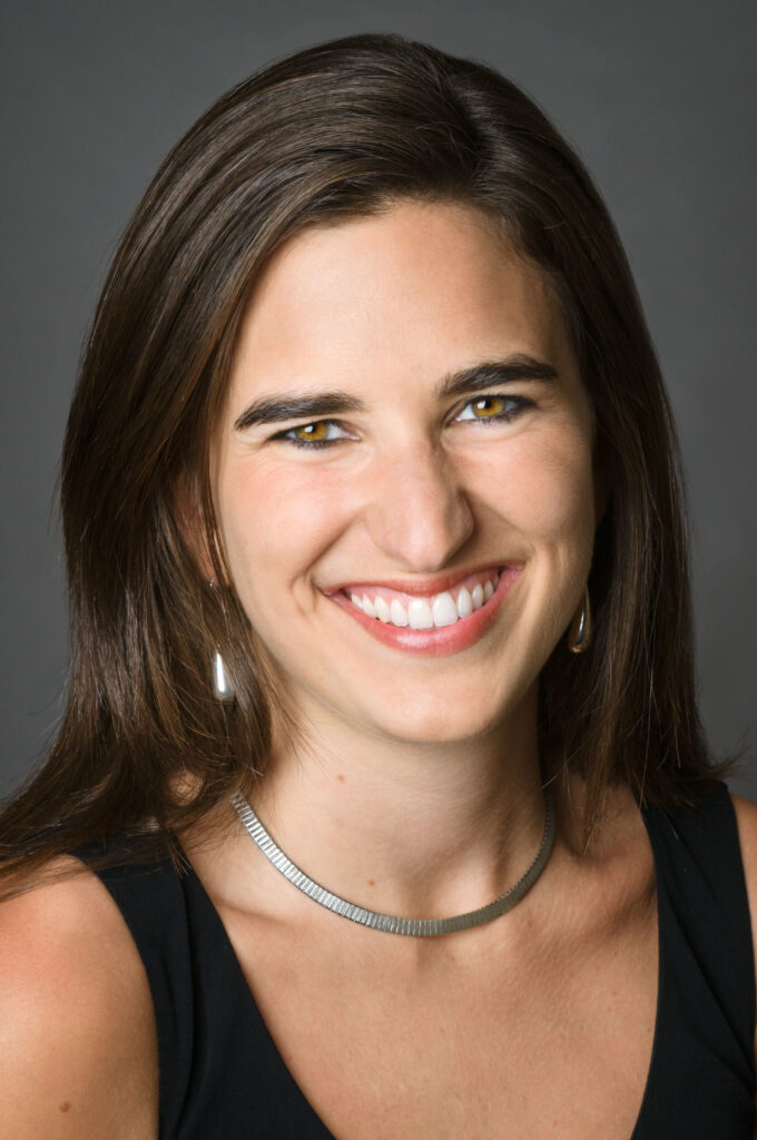 Headshot of a woman in her 20s who has heritage from United Kingdom and Iran, she has light skin tone and long straight dark brown hair with a left side part. She is wearing a black tank top, silver necklace and pearl drop earrings. She is smiling at the camera. 