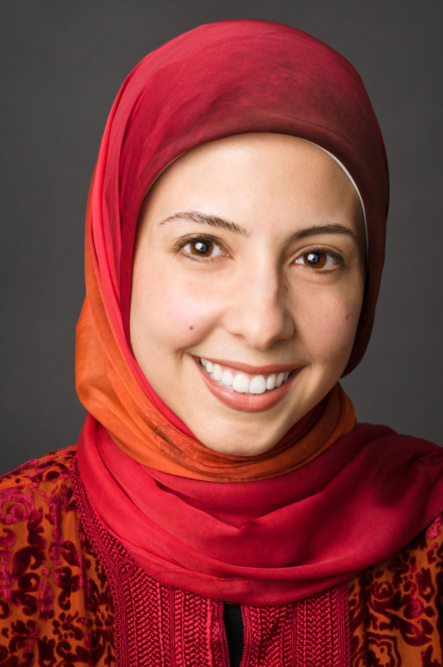 Headshot of a woman in her 20s who has heritage from Egypt with light skin tone. She is wearing a red to orange ombre hijab and a dark orange with velvet floral design top. She is smiling at the camera.