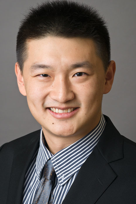 Headshot of a man in his 20s who has heritage from China with light skin tone and black short hair. He is wearing a black suit, white and blue striped shirt and a navy tie. He is smiling at the camera. 