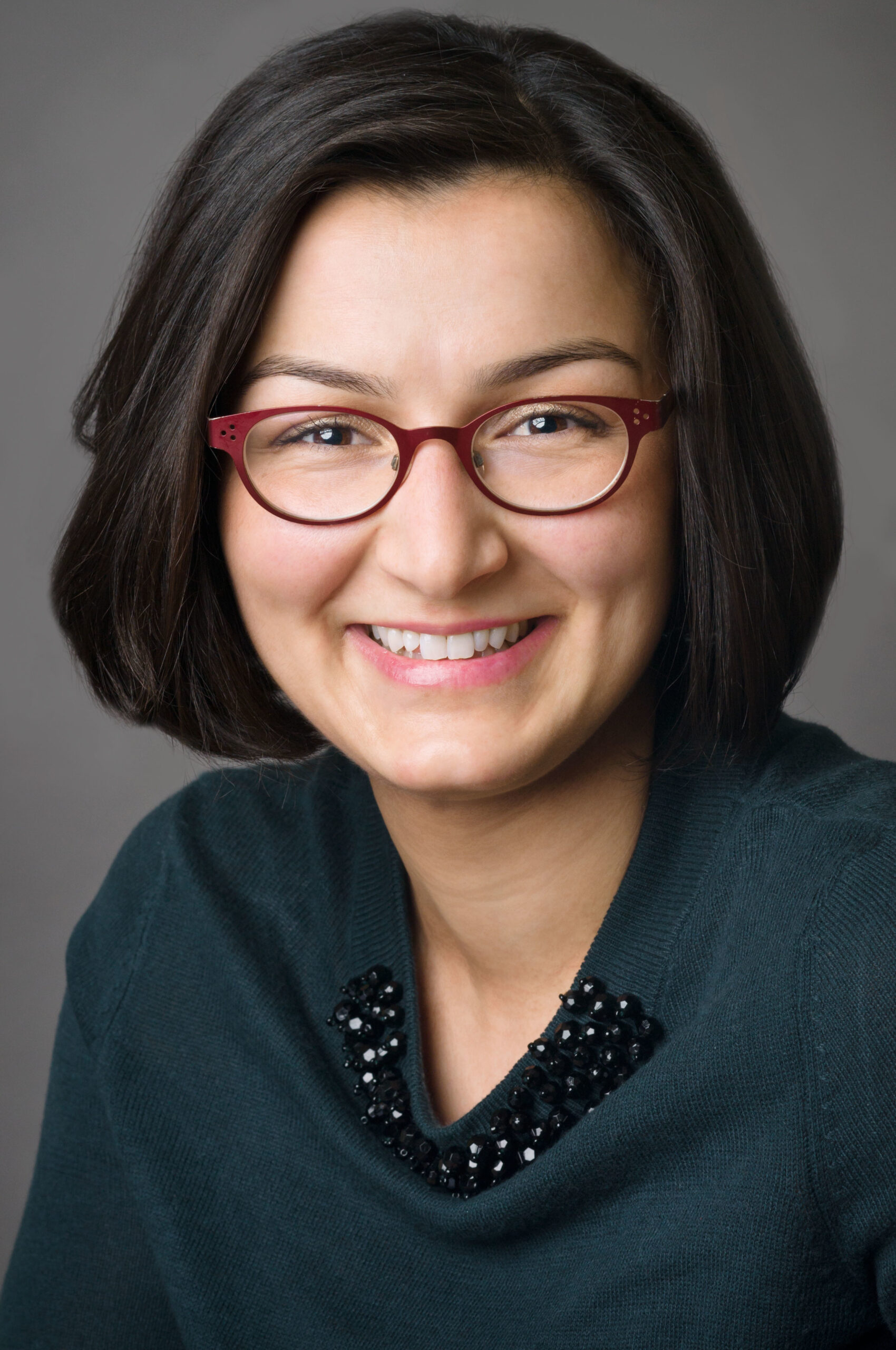 Headshot of a woman who has heritage from Iran with light skin tone and chin length black straight hair. She is wearing a dark teal top with a bejeweled flourish across the neckline and red rimmed oval glasses. She is smiling at the camera.