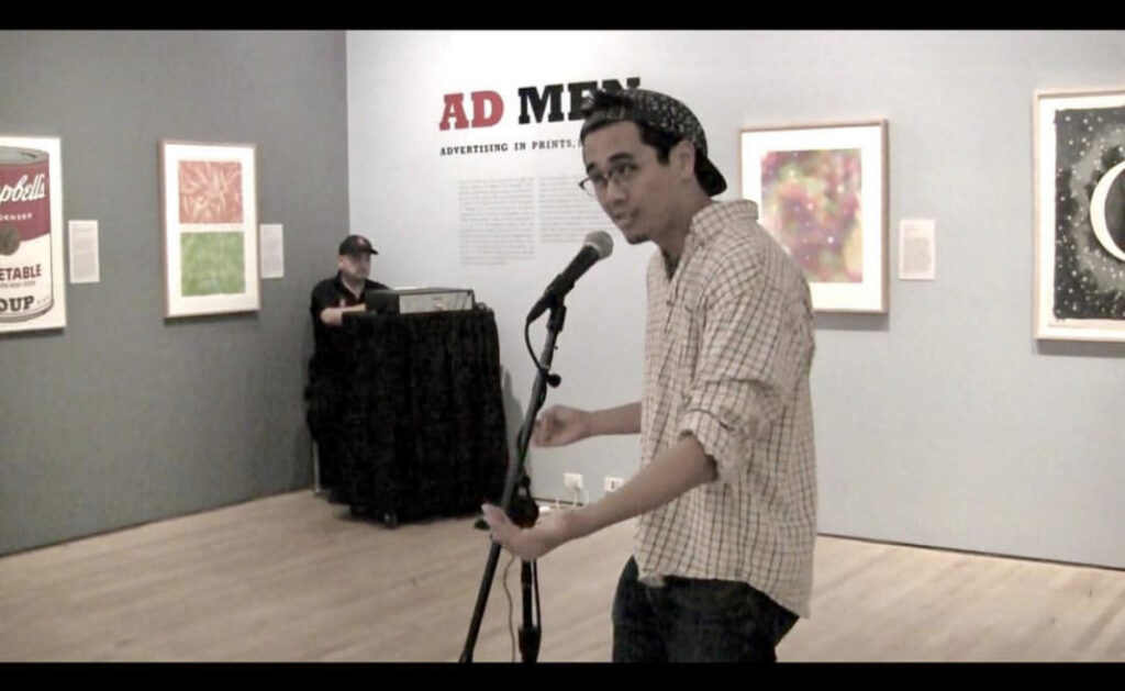 Photograph of a young man standing at a microphone in a gallery space. 